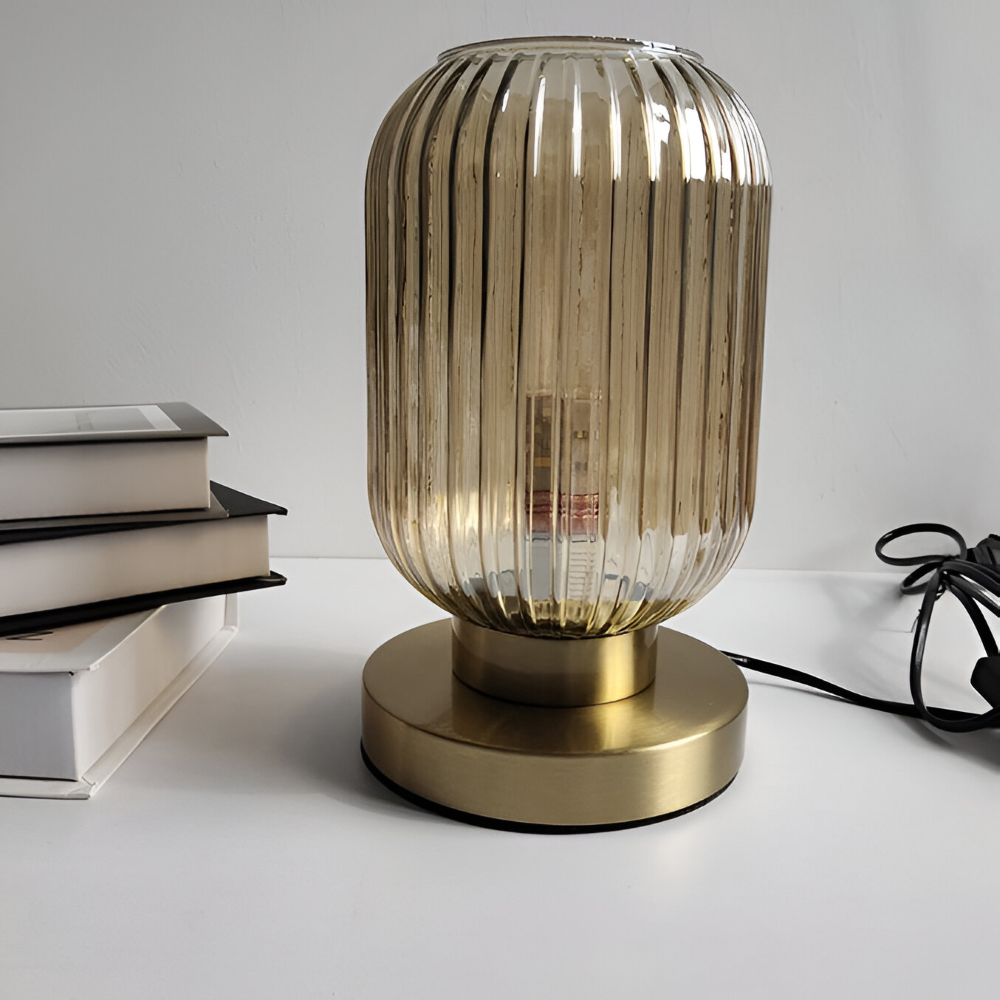 Szklana lampa stołowa na złotej podstawie obok książek