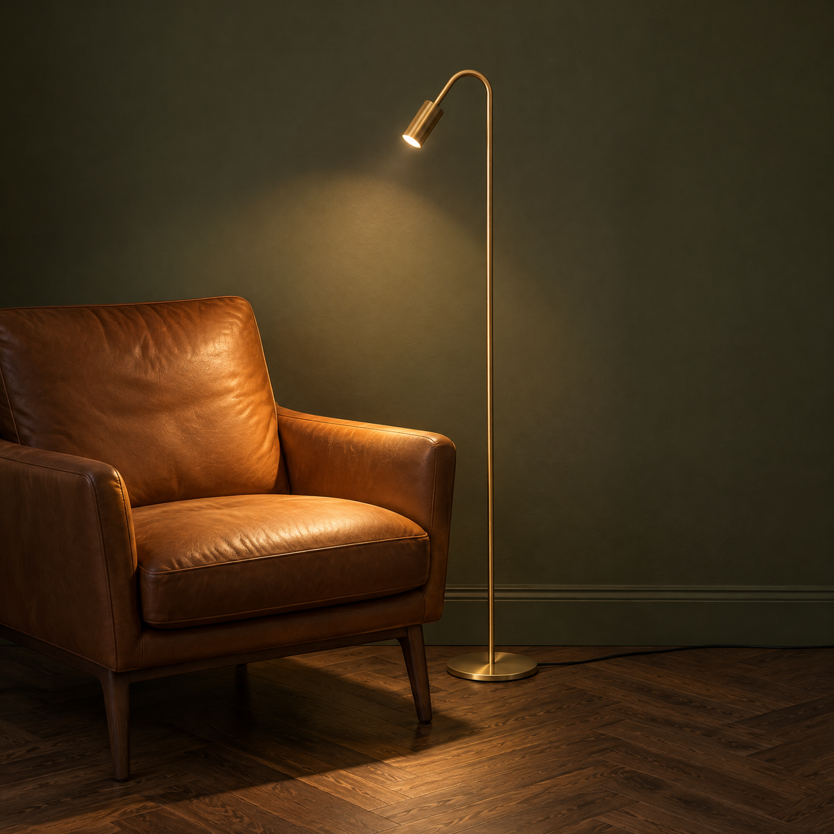 Wygięta lampa stojąca z miedzi w kolorze złotym jako lampa do czytania obok skórzanego fotela, włączona, z ciepłym snopem światła