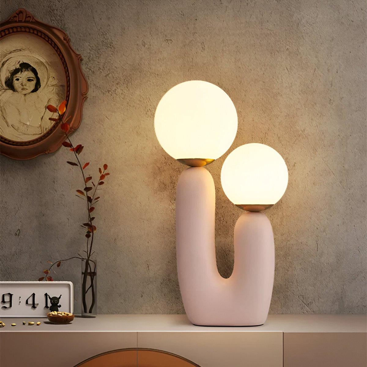Różowa lampa kaktus zapalona na komodzie obok zegara i suszonych gałązek